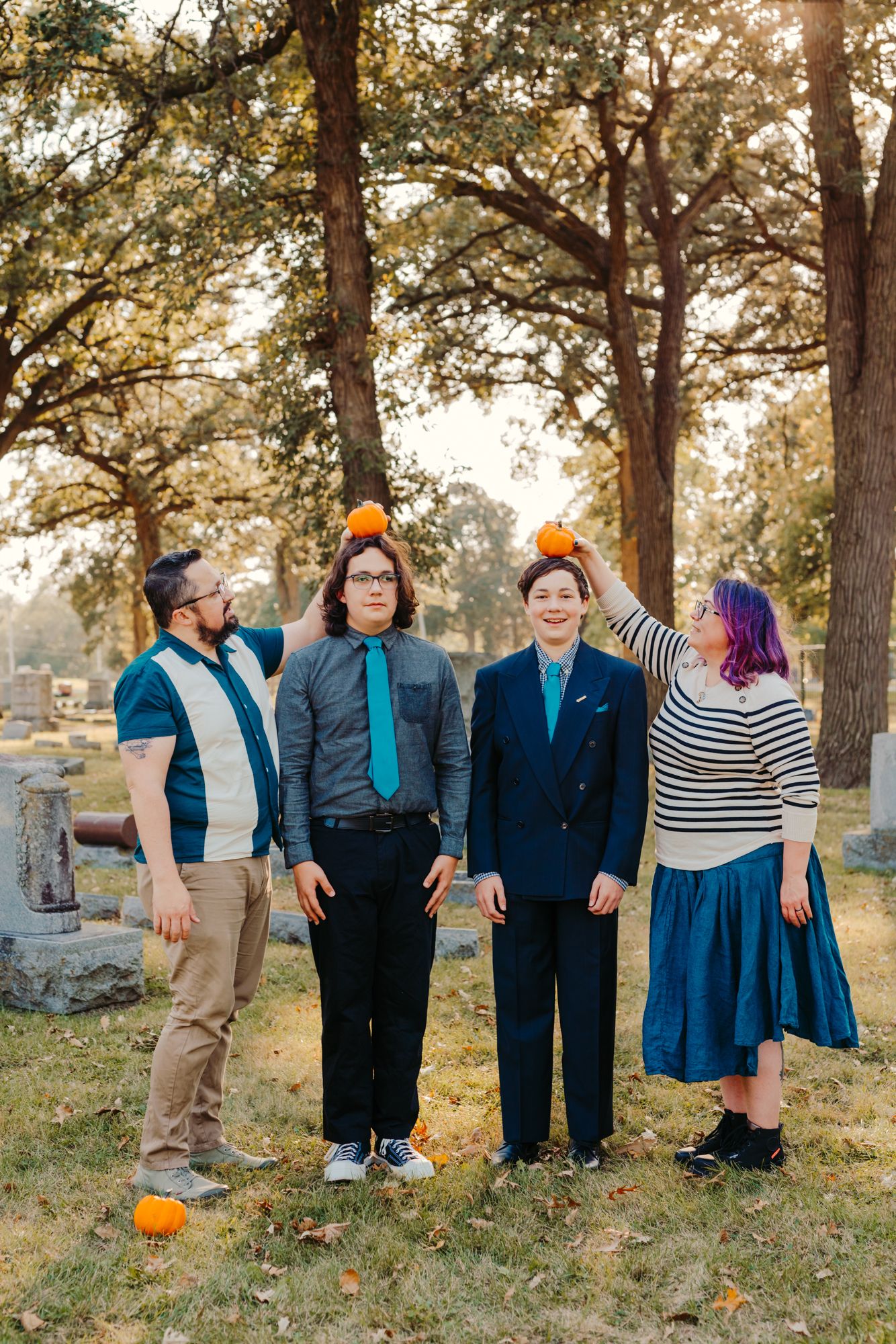 Four people in stylish outfits balance small pumpkins on their heads while posing in a cemetery during a playful autumn photo session.