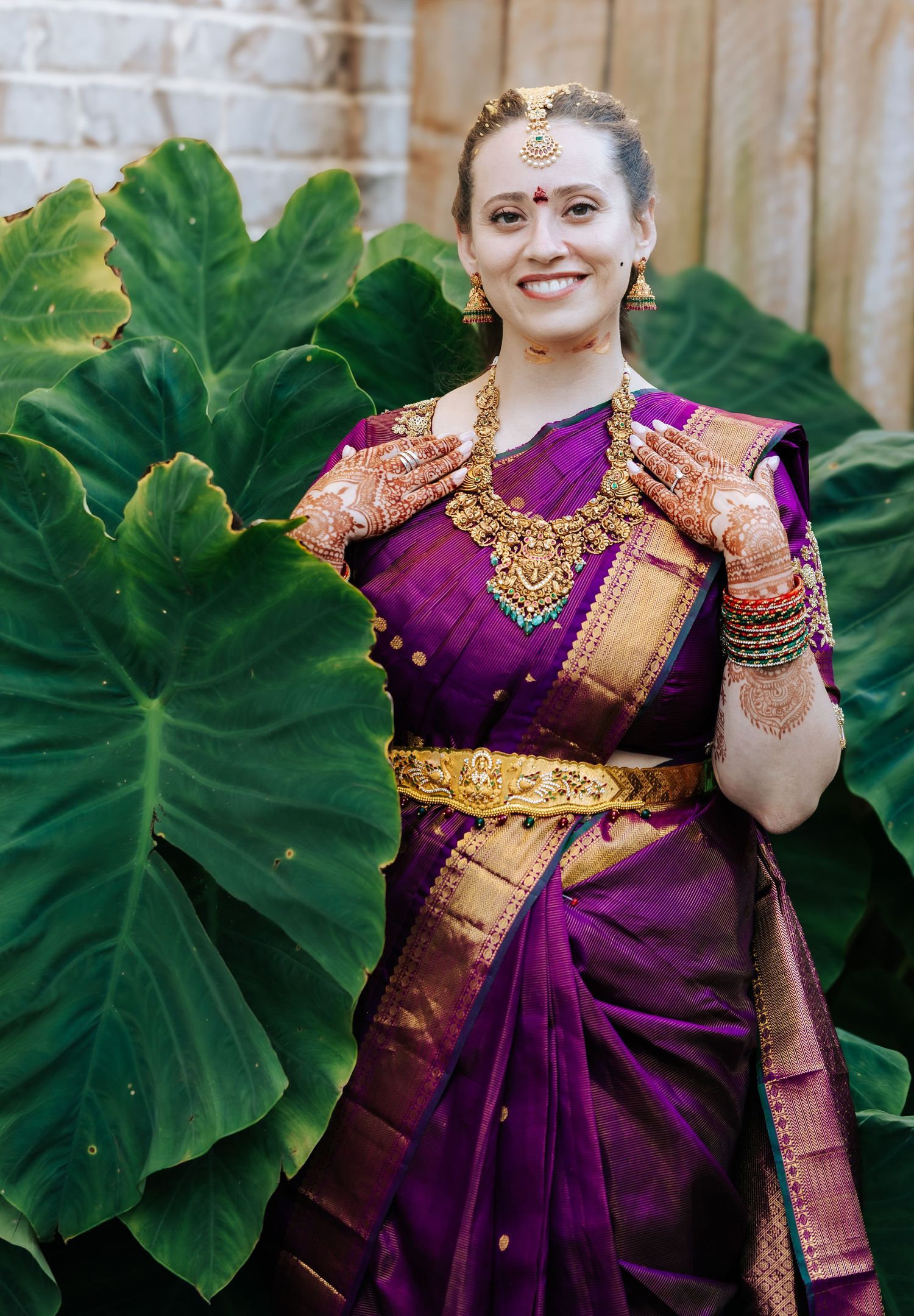 Smiling woman in a purple silk saree with gold border, temple jewelry and henna-decorated hands, standing before large green leaves.