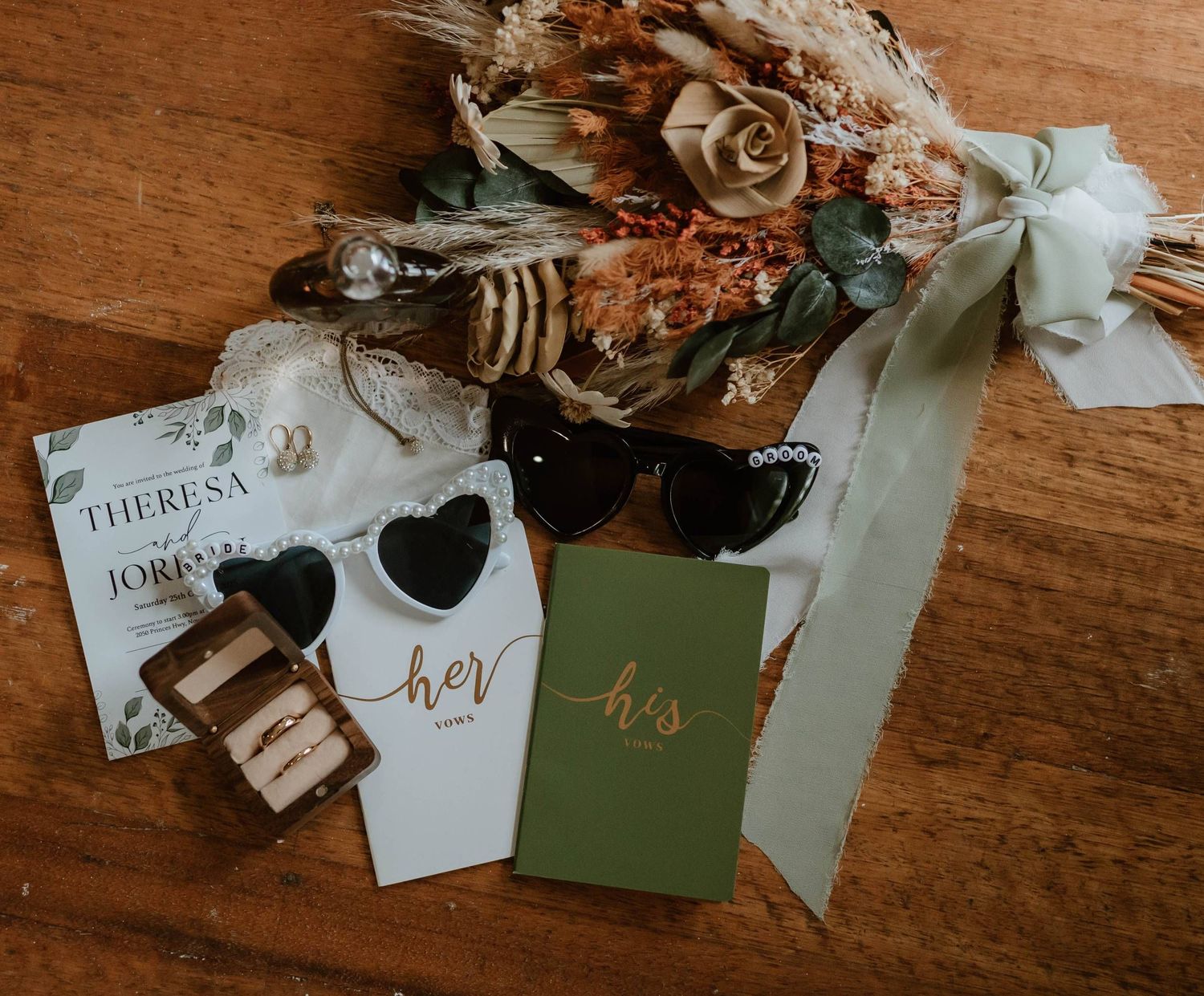 Wedding flat lay with dried floral bouquet, sunglasses, rings, and his and her vow books on wood.