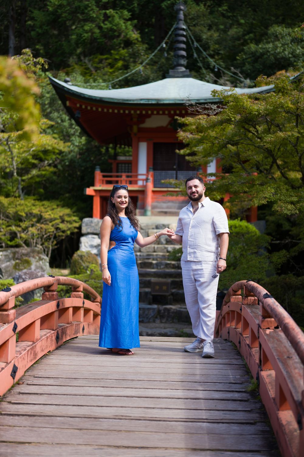 Couple holding hands on a red wooden bridge with a traditional Japanese pagoda and lush green trees in the background.