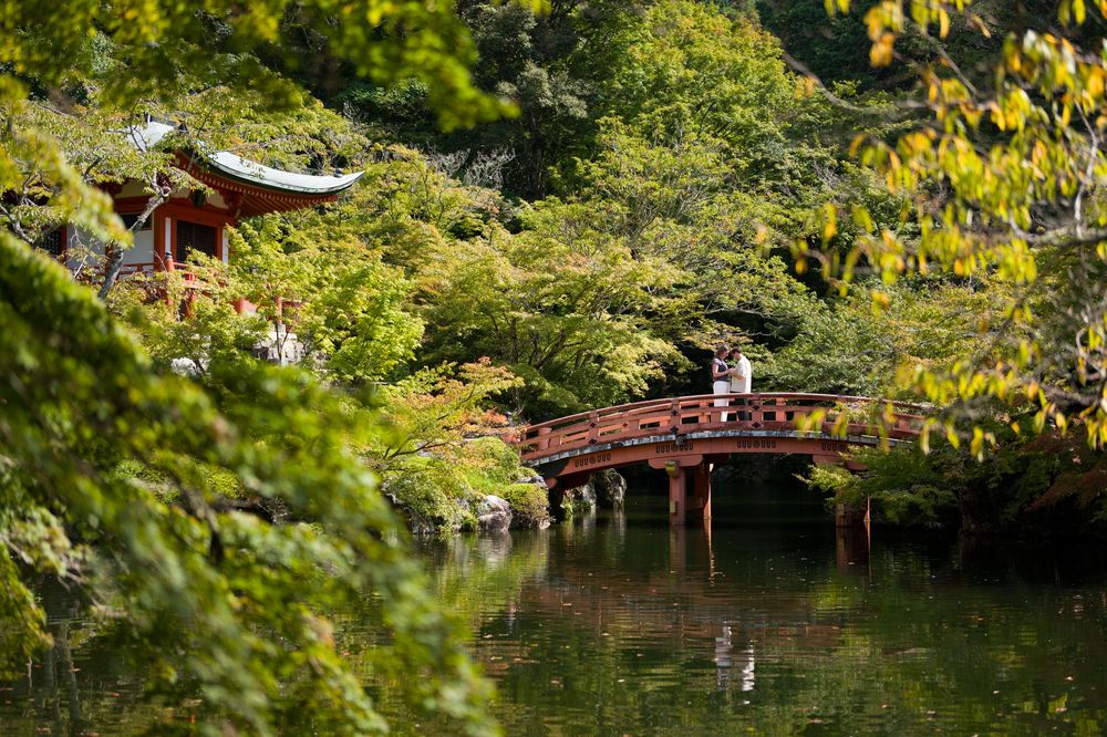 Red wooden arched bridge and pavilion with a curved roof surrounded by lush green trees reflecting in a calm pond at a Japanese garden.