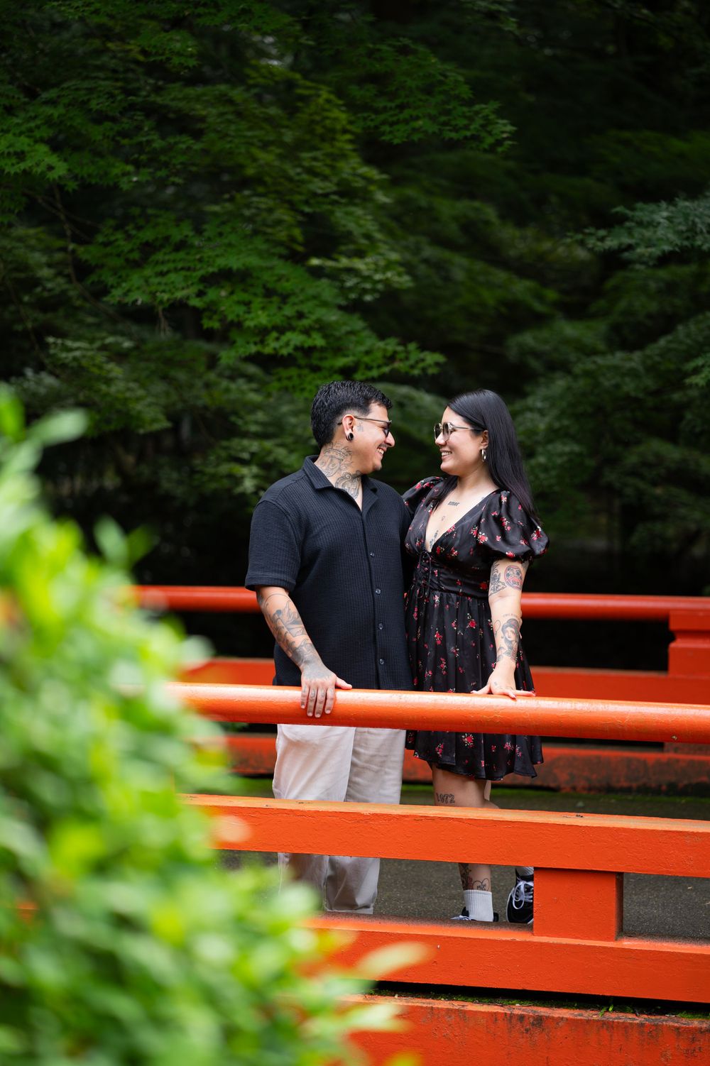 Smiling couple in matching dark outfits stand close beside a bright orange-red wooden railing with lush green trees in the background.