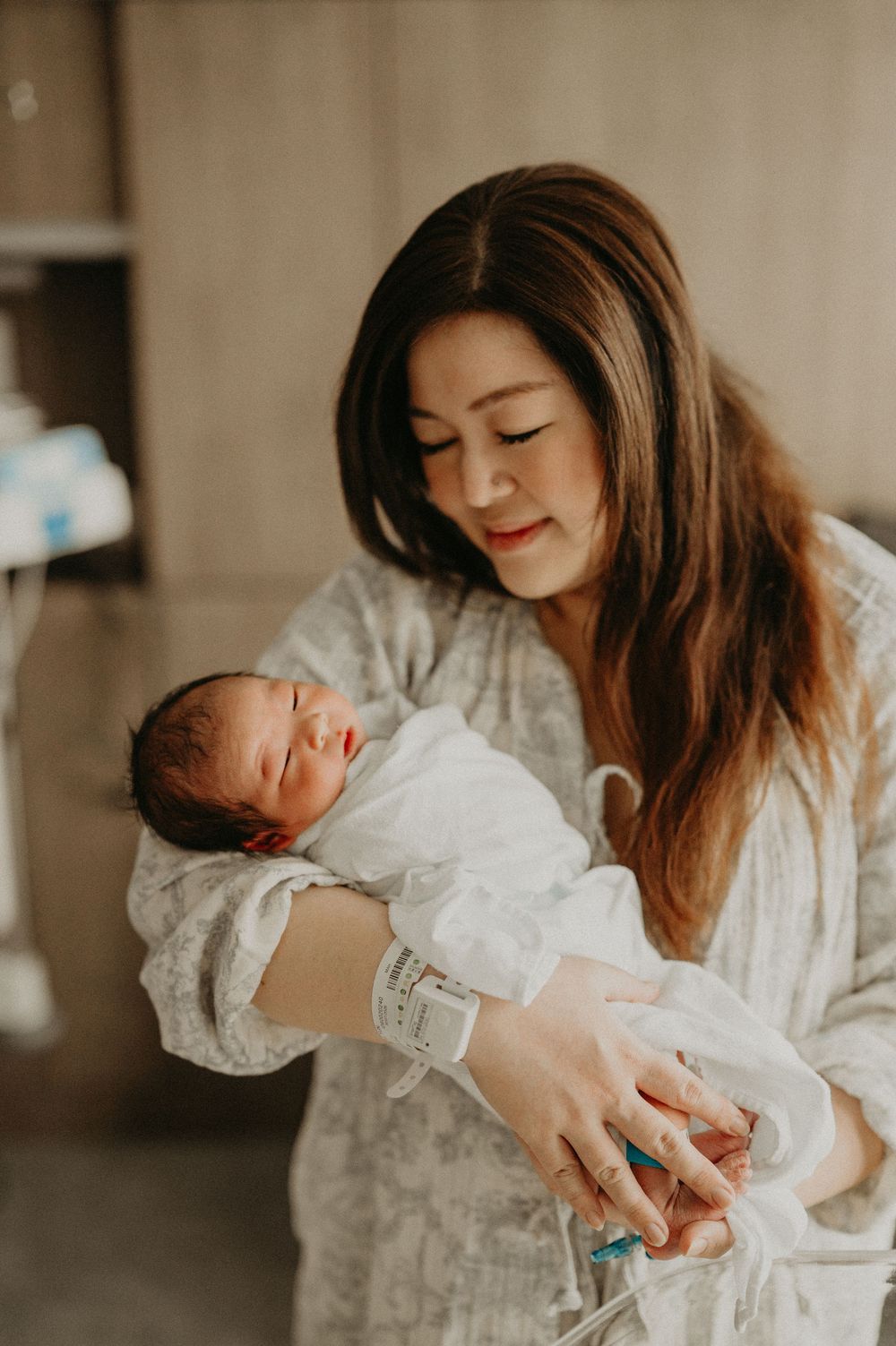 A smiling new mother in a patterned hospital gown and ID bracelet gently cradles her sleeping newborn baby wrapped in a white blanket.