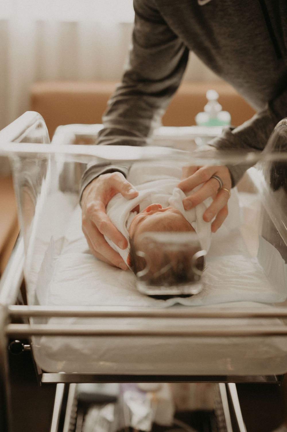 Parent gently tucks a swaddle blanket around a sleeping newborn baby in a clear hospital bassinet in a softly lit maternity room.