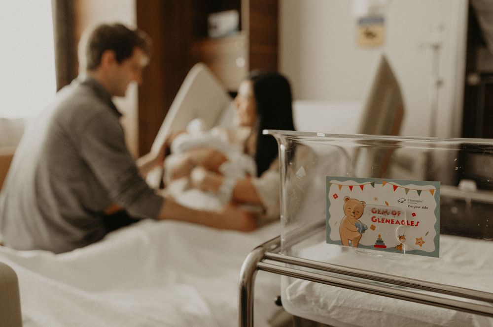 New parents sit on a hospital bed holding their swaddled newborn baby, with a clear bassinet and teddy bear name card in the foreground.
