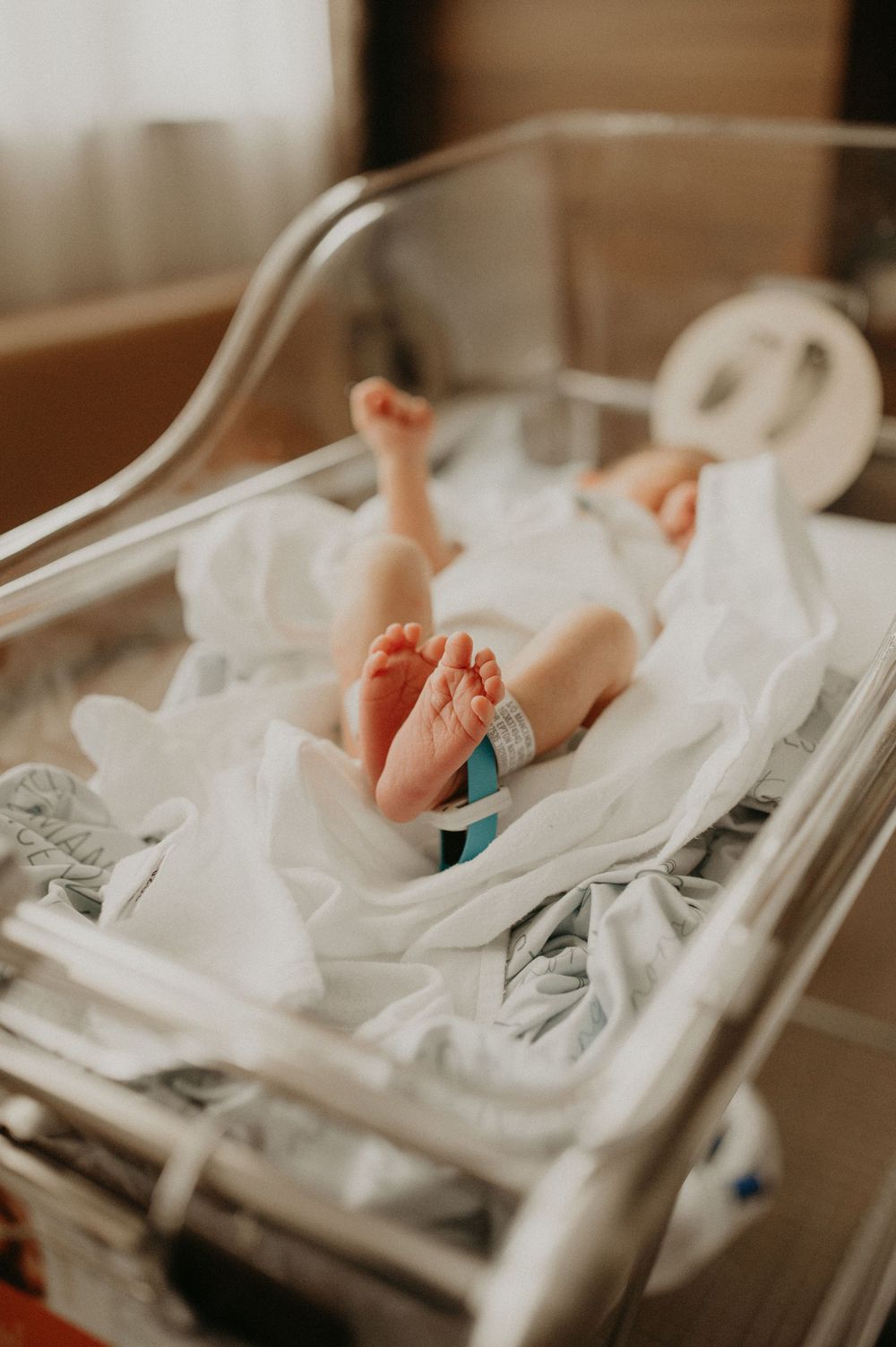 Newborn baby with a hospital ID bracelet on its ankle lying wrapped in white blankets inside a clear bassinet in a hospital room.