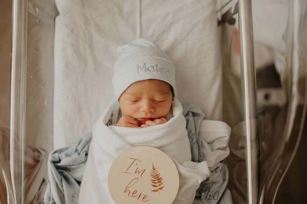 Sleeping newborn baby in a white knit hat swaddled in a blanket in a hospital bassinet beside a wooden "I'm here" birth announcement sign.