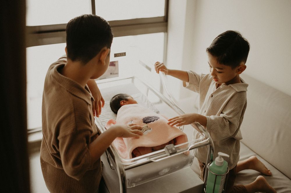 Two young brothers in matching beige outfits gently look at their newborn sibling sleeping in a hospital bassinet.