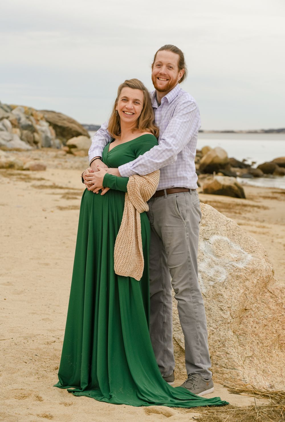A smiling couple embraces on a sandy beach with rocks and water behind them, she in a flowing green gown, he in a button-up shirt.