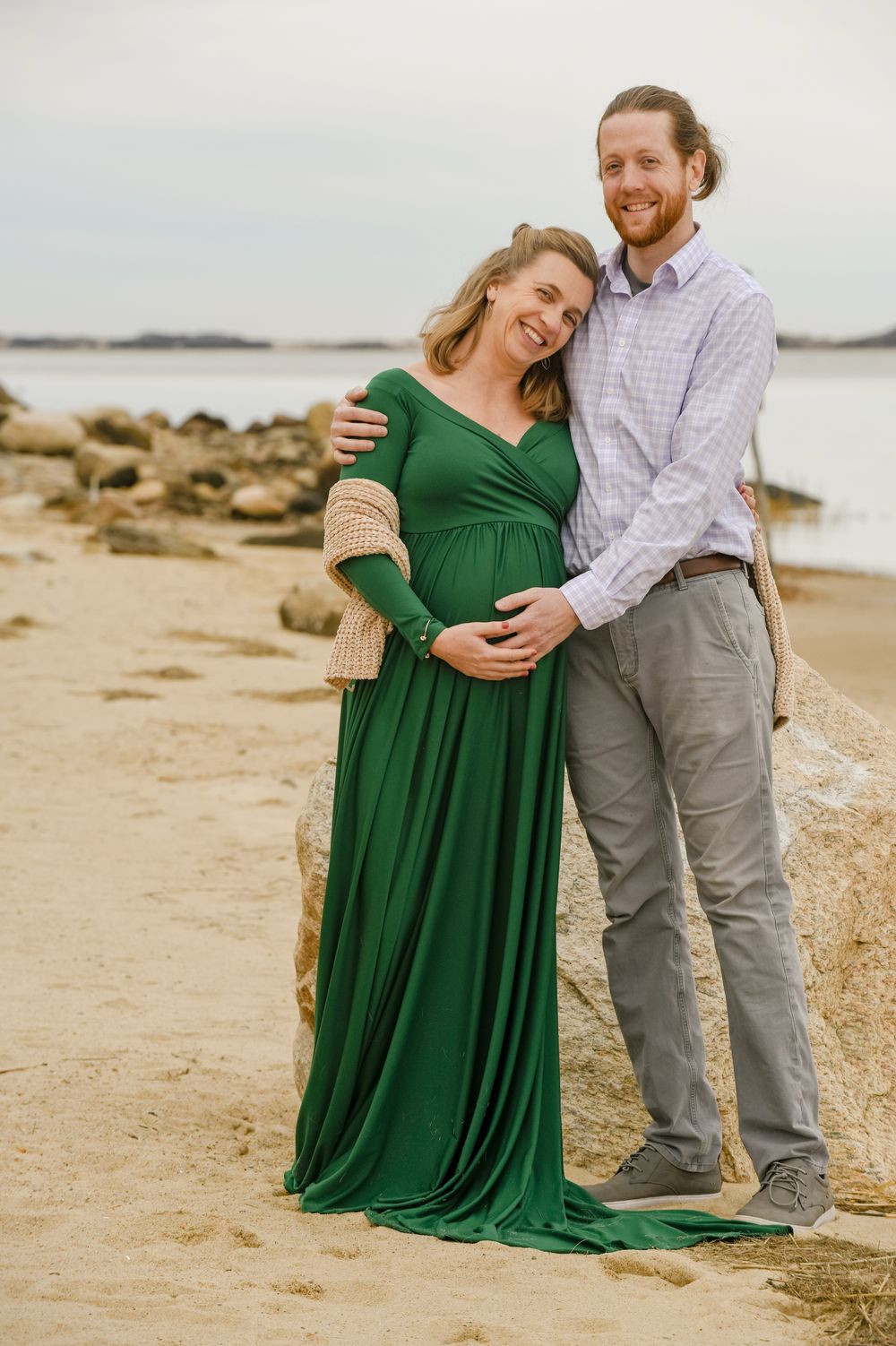 Smiling expectant couple embracing on a sandy beach, the woman in a green maternity gown cradling her baby bump beside rocks and calm water.