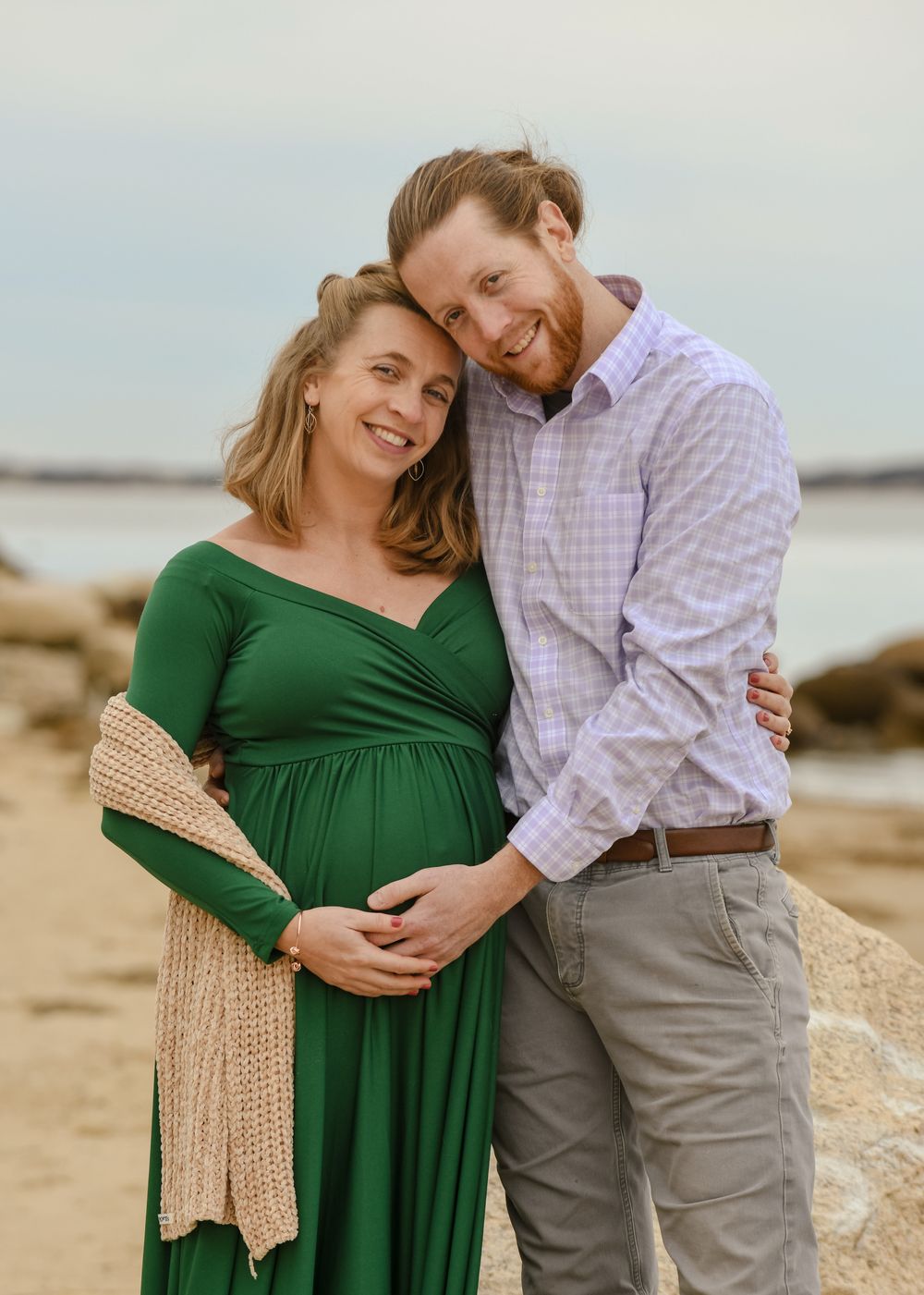 Smiling expecting couple embracing on a rocky beach, the woman in a green maternity dress cradling her baby bump beside her bearded partner.