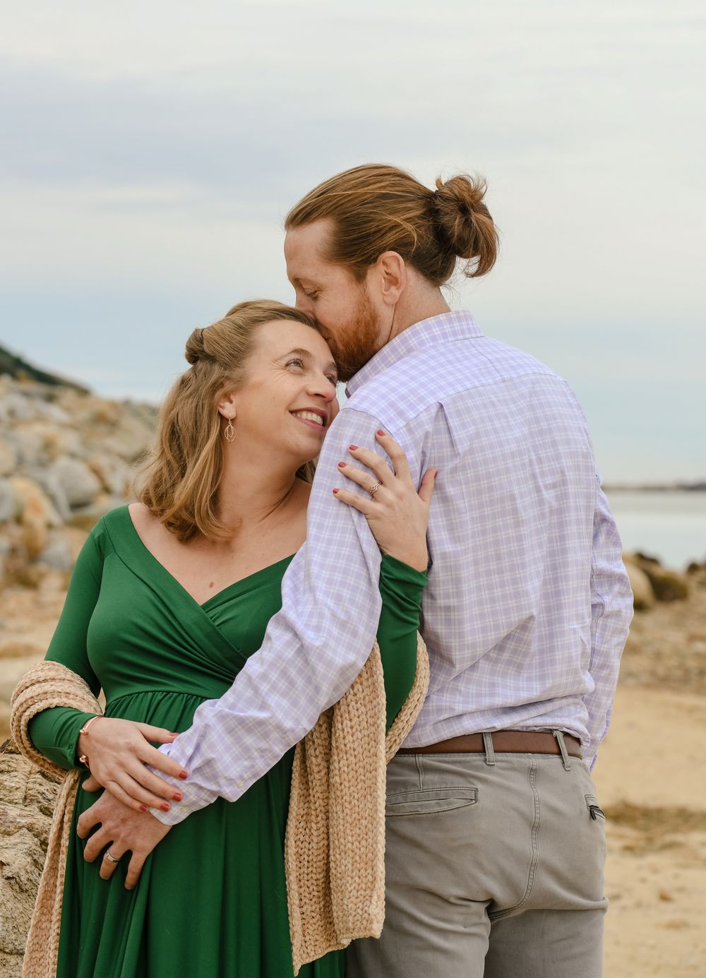 A smiling pregnant woman in a green dress embraces her bearded partner, who kisses her forehead, on a rocky beach at sunset.