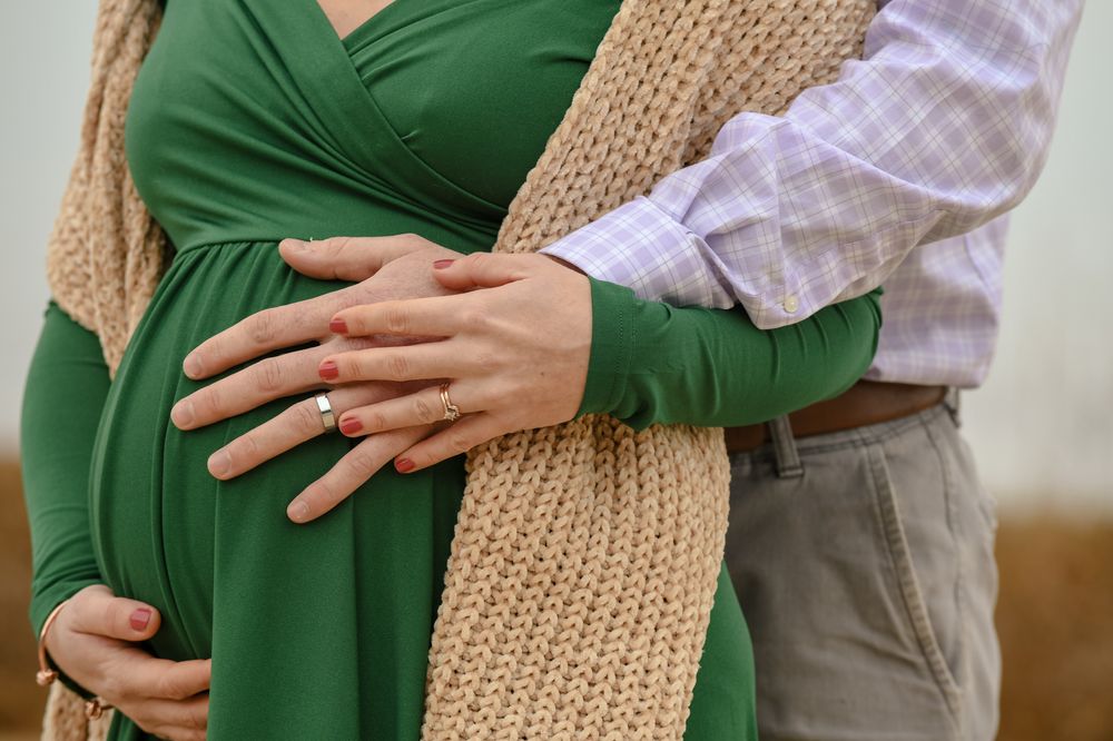 Couple embracing in a maternity photo, hands with wedding rings resting on the pregnant belly of a woman in a green dress and knit scarf.