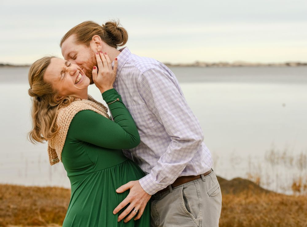 Smiling pregnant woman in a green dress laughs as her partner kisses her cheek while they embrace by a calm lake at sunset.