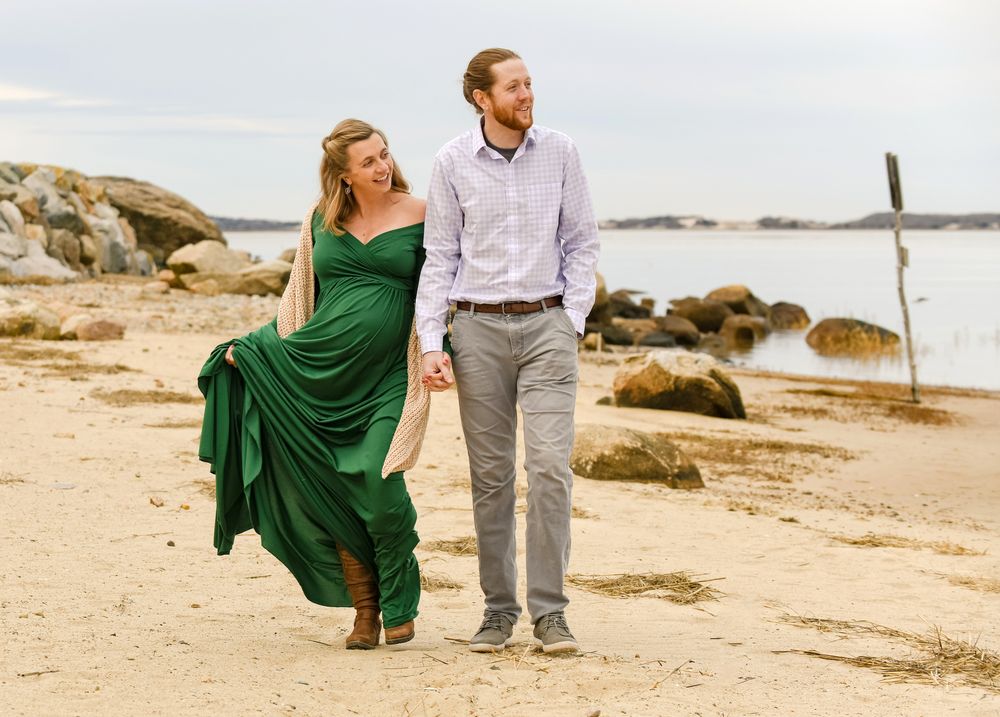 Smiling couple holding hands and walking along a rocky sandy beach, she in a flowing green dress and he in a light blue shirt.