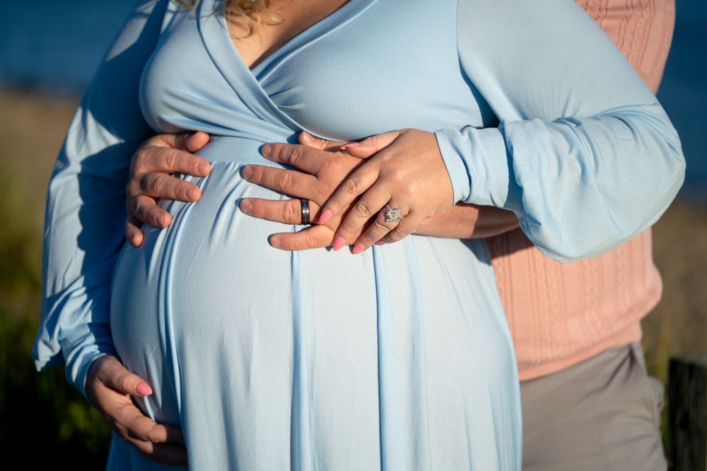 Pregnant woman in a light blue wrap dress cradling her baby bump as her partner's hands rest on top, showing wedding rings, outdoors.