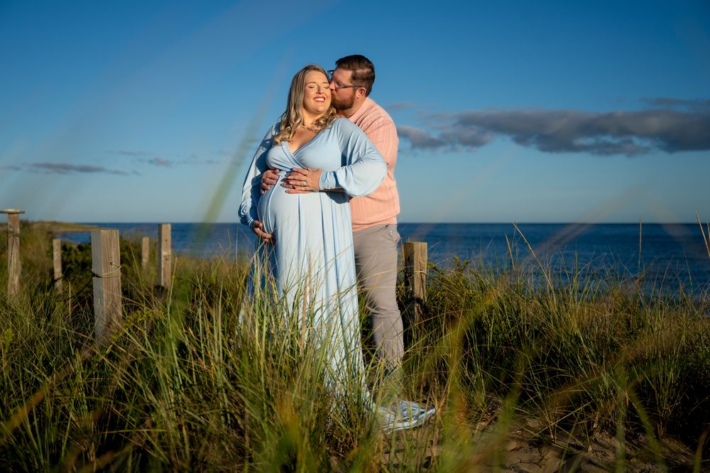Expecting couple embracing in beach grass at sunset, the pregnant woman in a flowing blue gown as her partner kisses her cheek by the ocean.