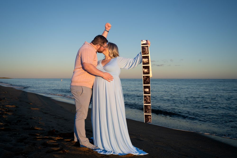 Expecting couple embracing and kissing on a sunset beach while holding a vertical strip of ultrasound photos beside the ocean.