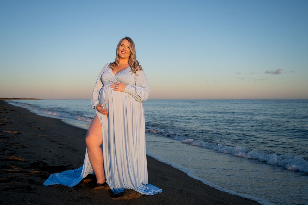 Pregnant woman in a flowing pale blue maternity gown cradling her baby bump on a sandy beach at sunset beside gentle ocean waves.