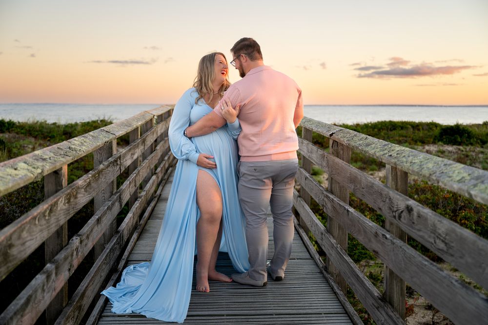 Expecting couple embracing on a wooden boardwalk at sunset, the woman in a flowing blue maternity gown, with ocean and dunes behind them.