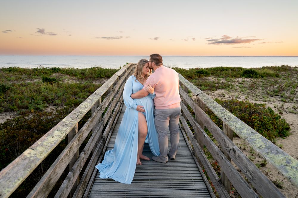 Expecting couple embracing on a wooden beach boardwalk at sunset, the woman in a flowing blue maternity gown, with dunes and ocean behind.