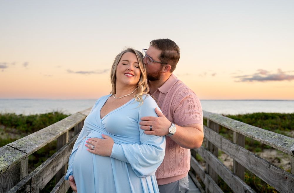 Smiling pregnant woman in a light blue dress and a man kissing her cheek pose on a wooden beach boardwalk at sunset.