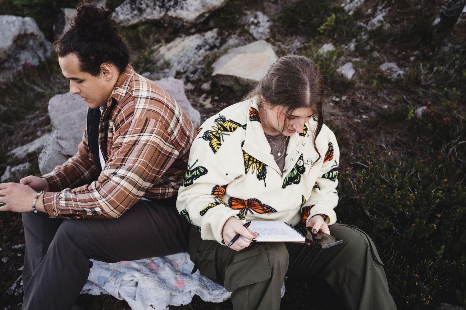 Two people sit back to back on a rocky hillside, one in a plaid flannel shirt, the other in a butterfly-print jacket writing in a notebook.