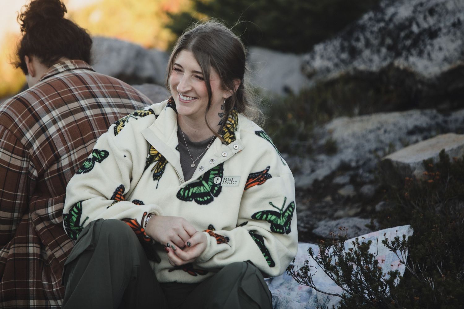 Smiling woman in a cream fleece jacket with butterfly patterns sits outdoors on rocky terrain beside a person in a plaid flannel shirt.