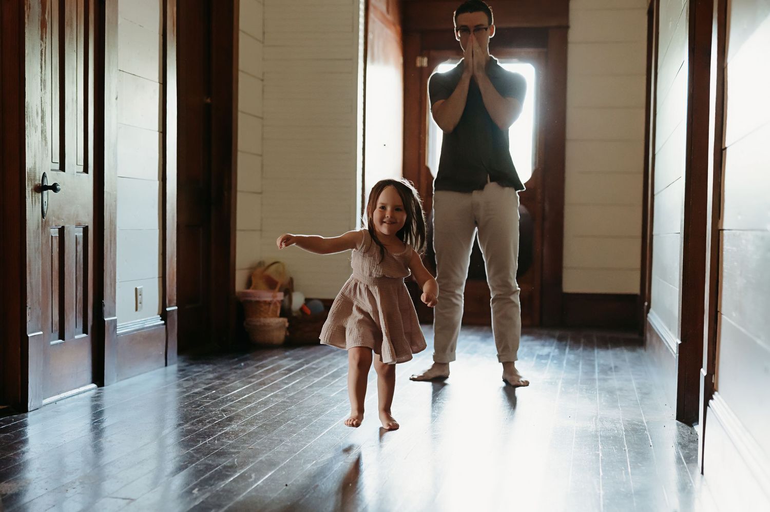 A smiling toddler girl in a dress runs barefoot down a sunlit wooden hallway toward a man standing with his hands clasped near his face.
