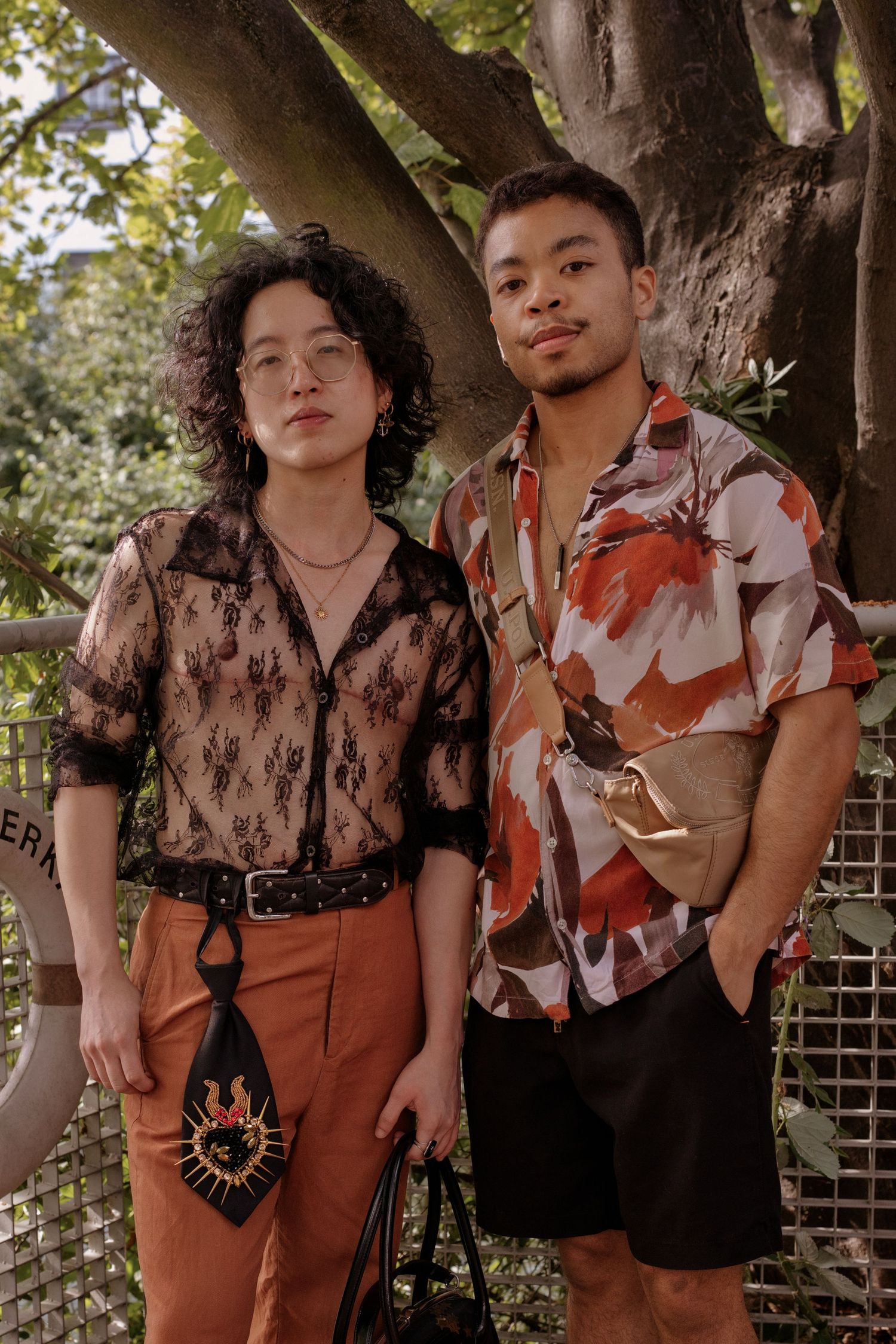Two stylish young people pose outdoors near a tree, wearing trendy outfits with accessories.