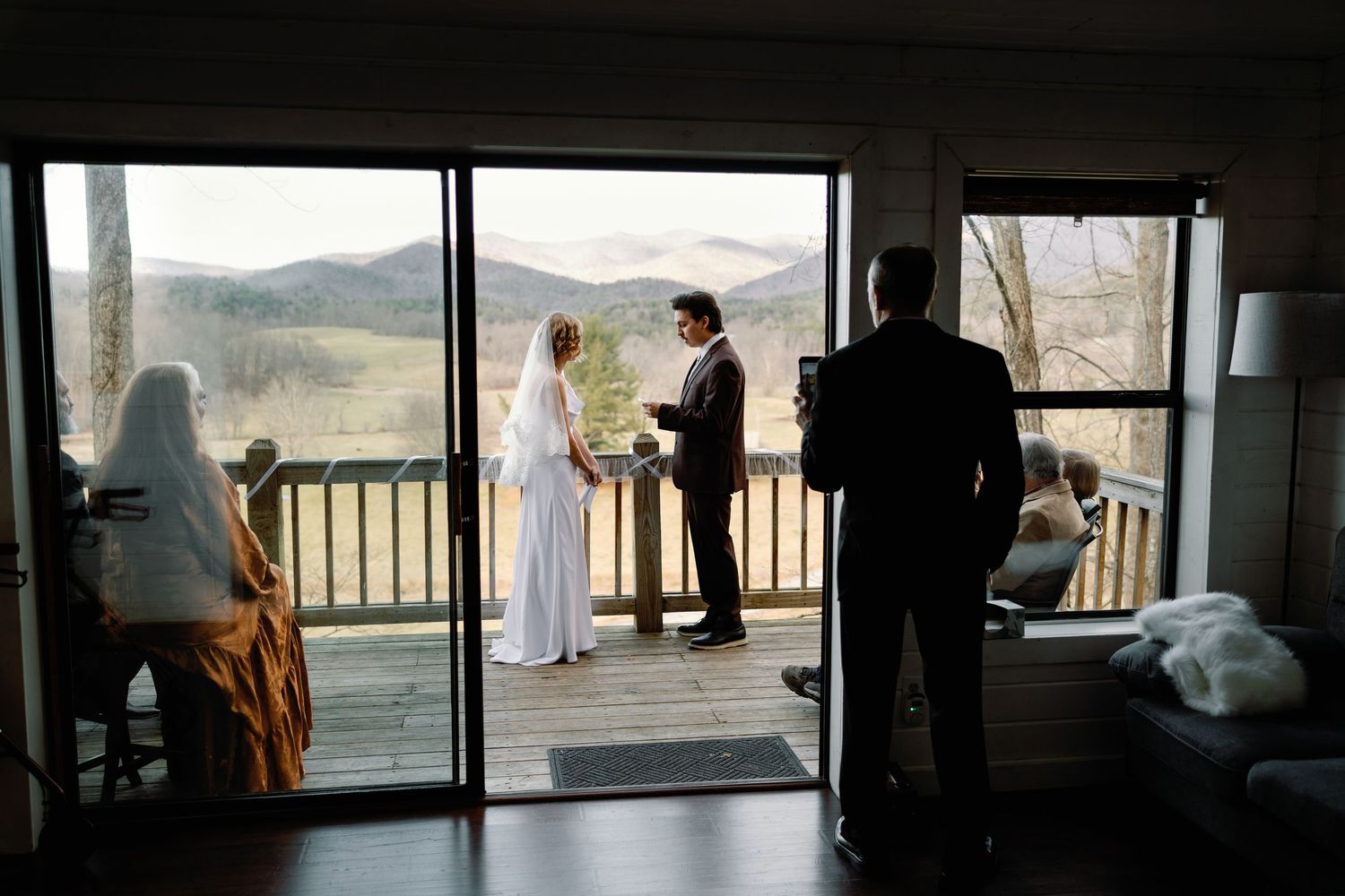 Couple exchanging vows on a mountain cabin deck, viewed through glass doors with scenic hills in background.