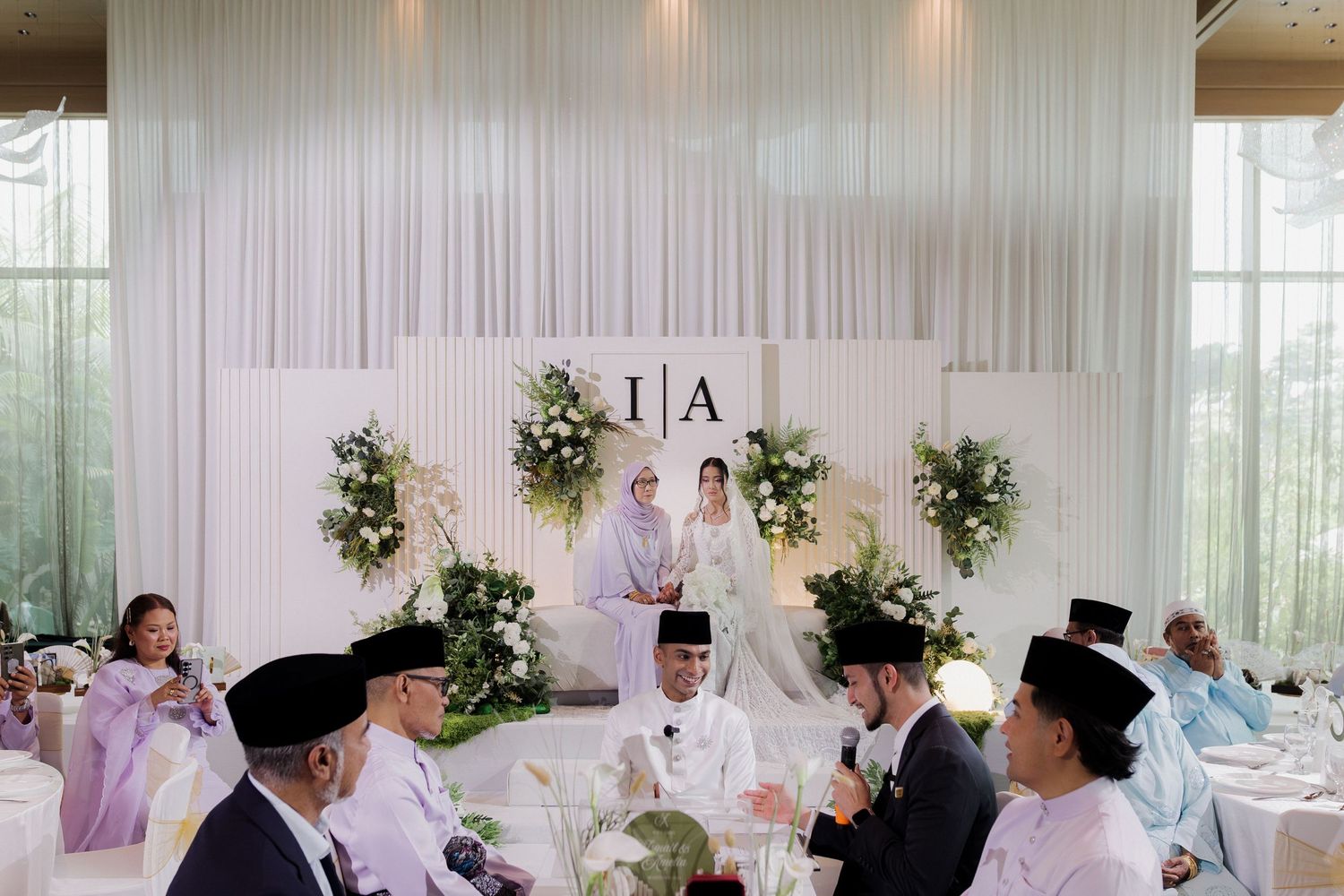 Muslim couple in traditional Javanese wedding attire sitting together at an outdoor ceremony with floral decorations and ocean view.