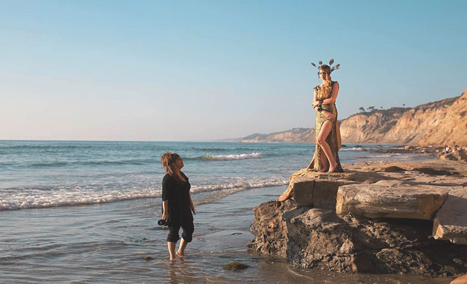 Photoshoot at a rocky beach at golden hour, one in a gold gown and crown standing on the rocks