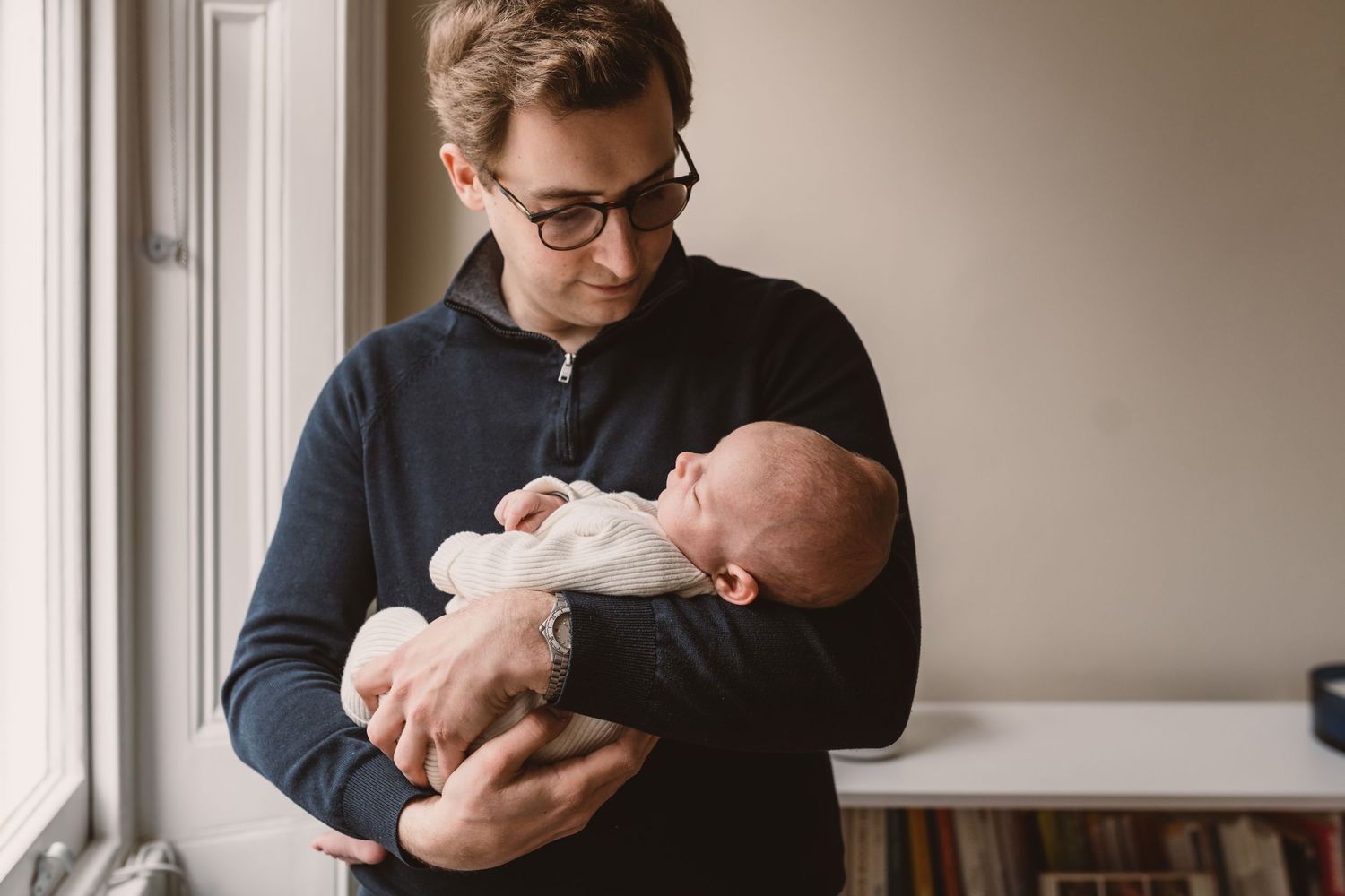 A father wearing glasses and a navy sweater tenderly holds his newborn baby near a bright window.