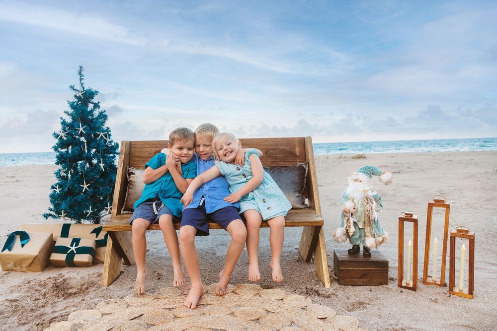 Three young children in blue hug on a wooden beach bench near a Christmas tree, a Santa figure and wooden lanterns.