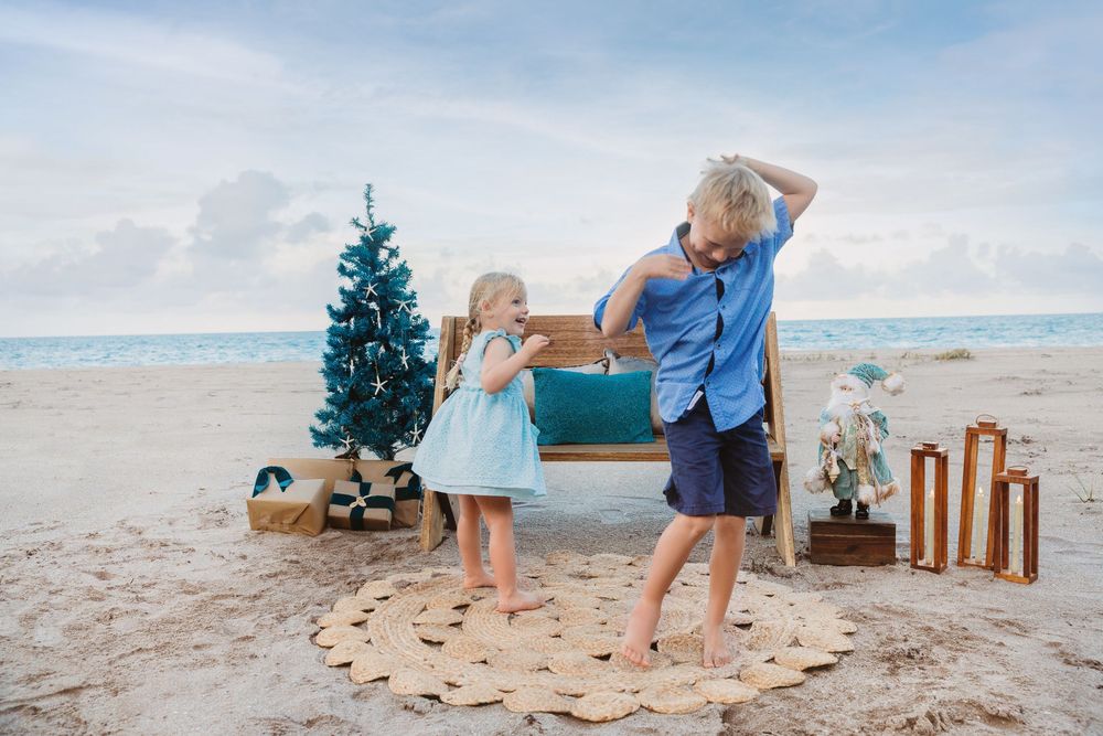 Two blonde children play on a sandy beach beside a small Christmas tree, a wooden bench, and festive decorations under a soft cloudy sky.