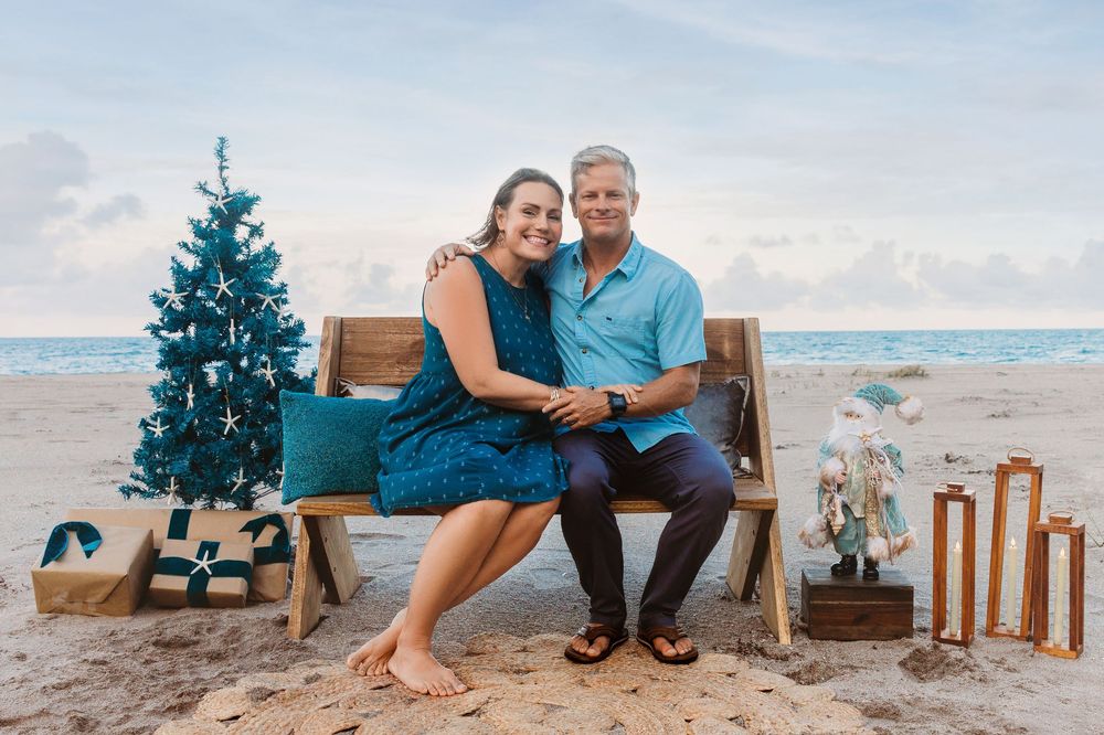 Smiling couple sits on a wooden bench on a beach beside a turquoise Christmas tree, wrapped gifts, a Santa figure and lanterns at sunset.