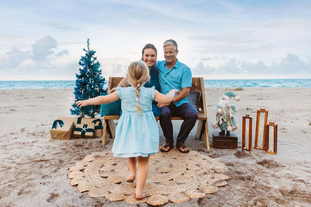 Smiling couple sits on a wooden bench on a beach while a little girl in a blue dress walks toward them near a Christmas tree and gifts.