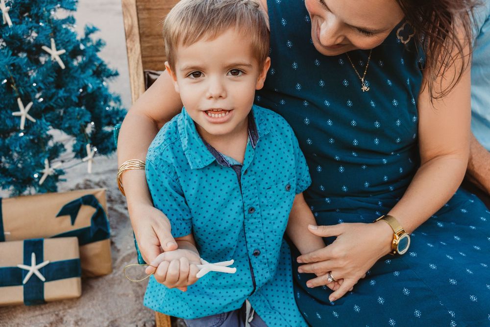 Smiling young boy in a teal button-up shirt sits beside his mother in a matching dress near a decorated Christmas tree and gifts.