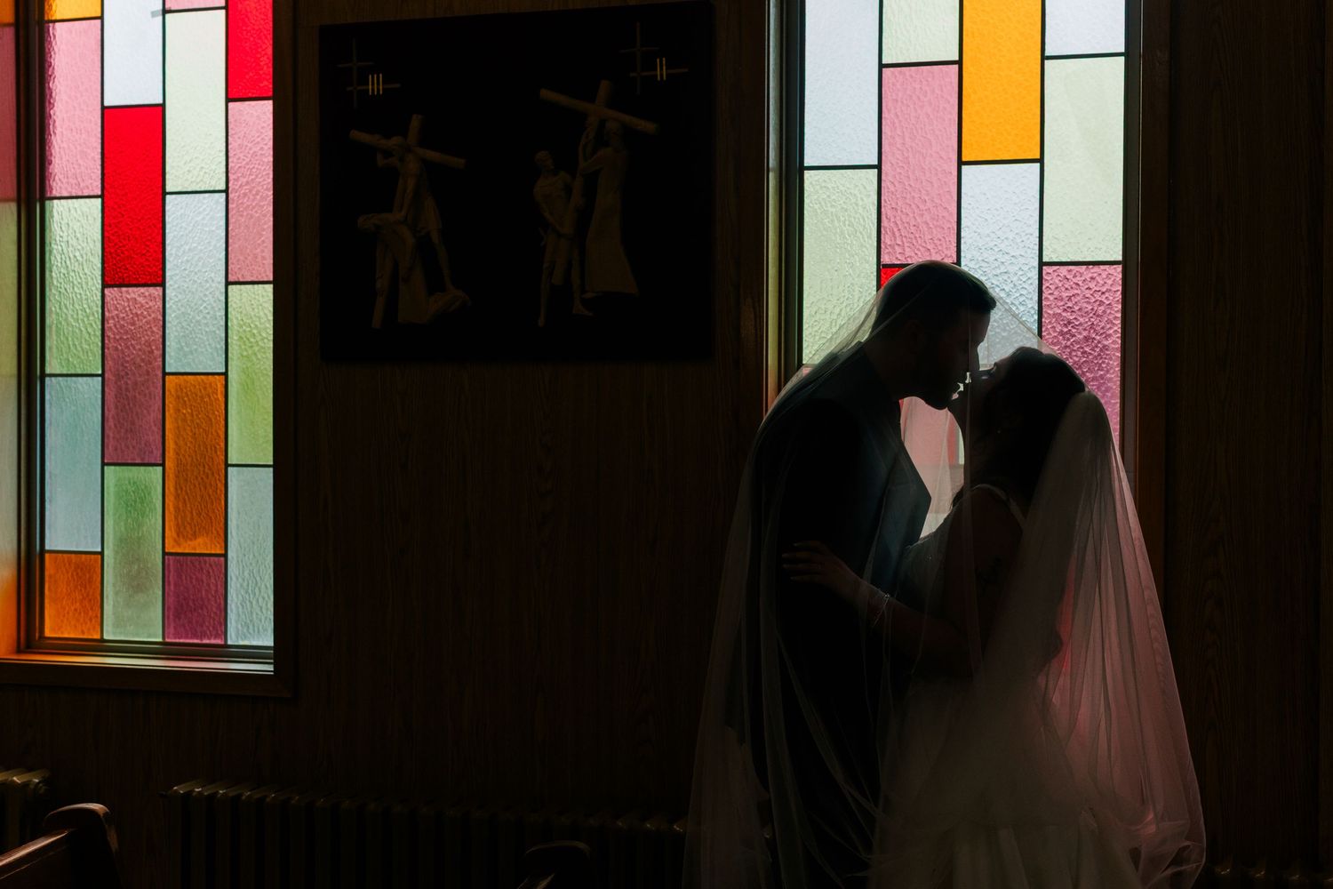 Silhouetted bride in a veil and groom share an intimate moment beside colorful stained glass windows in a dimly lit chapel.