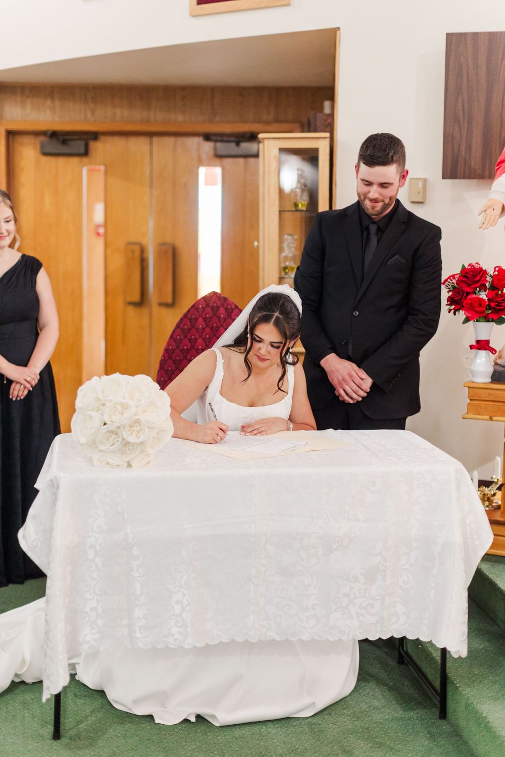 Bride in a white gown and veil signs the marriage register at a draped table as the groom in a black suit and a bridesmaid look on.