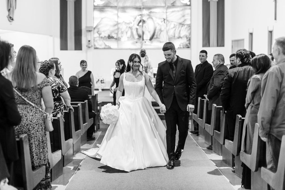 Bride in a white gown and groom in a dark suit walk hand in hand down the church aisle past smiling wedding guests in black and white.