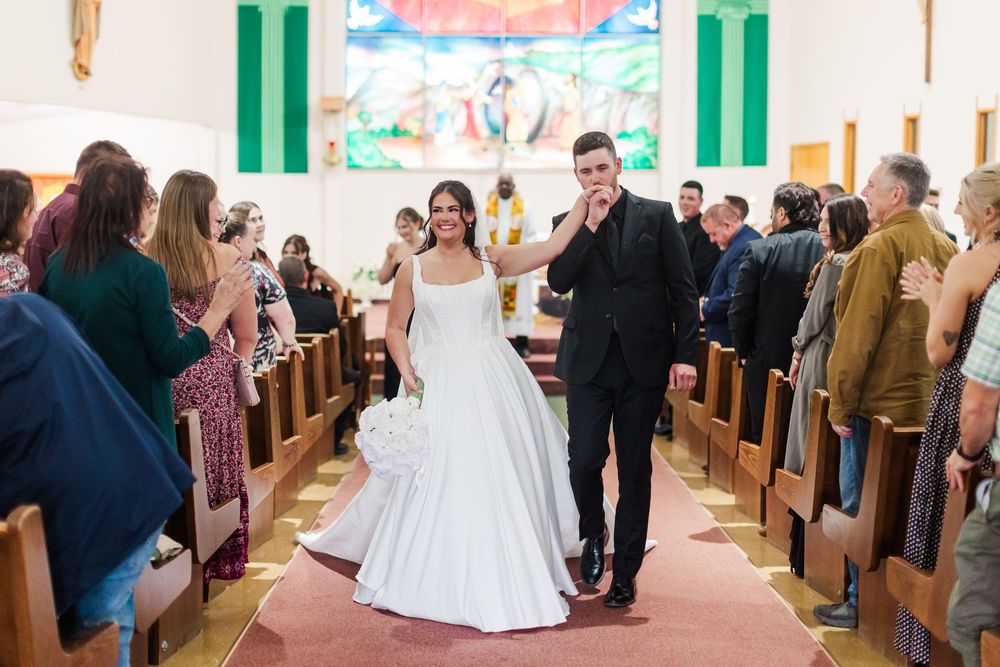 Bride in a white gown and groom in a black suit walk hand in hand down the church aisle as smiling wedding guests watch.