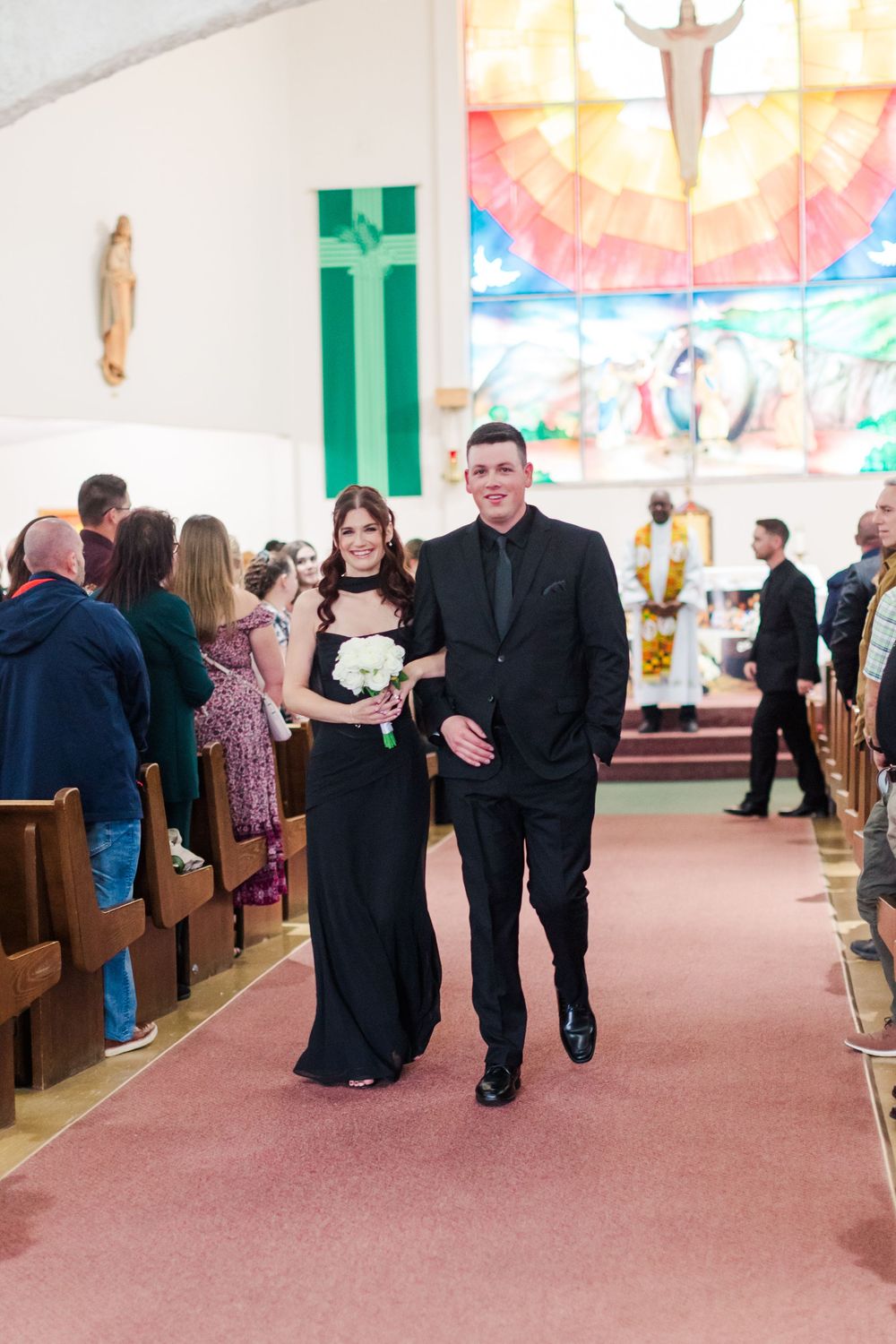 Bridesmaid in a black dress holding a white bouquet walks down the church aisle with a groomsman in a black suit during a wedding ceremony.