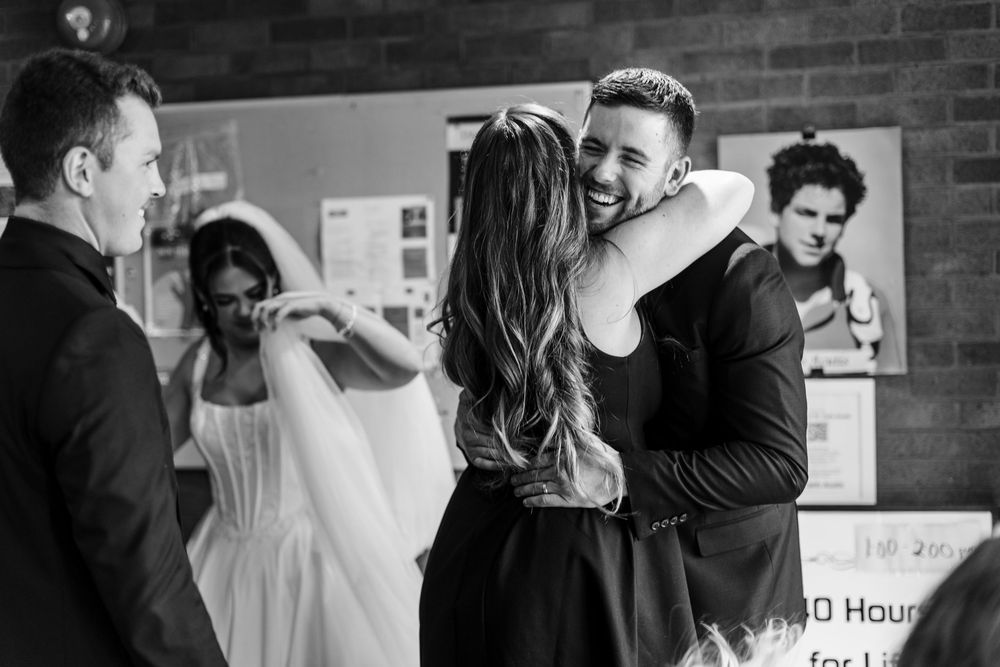 Black and white photo of a smiling man embracing a woman with long hair while a bride in a veil and gown stands nearby at a wedding.