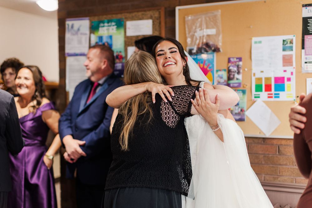 Smiling bride in a white wedding dress hugs a woman in black while guests look on in a room with bulletin boards.