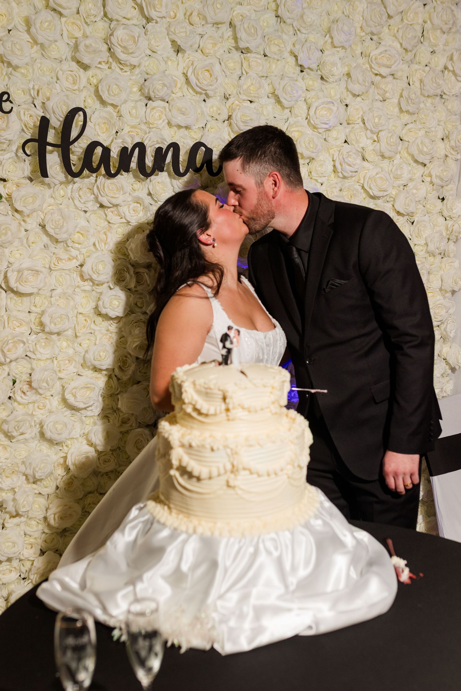 Bride and groom kiss beside a two-tier white wedding cake in front of a cream rose flower wall with a Hanna name sign.