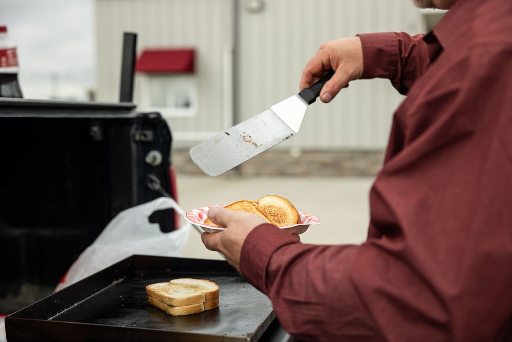 Person in a maroon shirt holds a metal spatula and a plated sandwich beside a black griddle with a slice of toast cooking outdoors.
