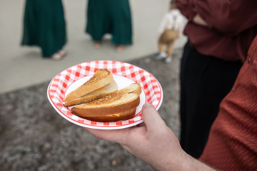 A hand holds a red and white checkered paper plate with two slices of toasted bread while people stand outdoors in the background.