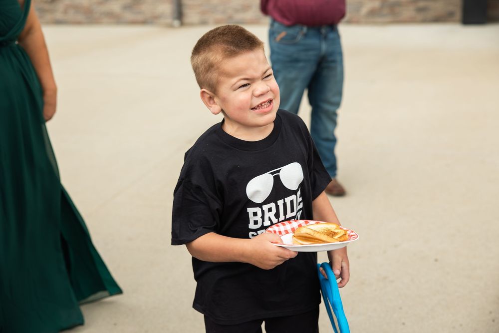 Smiling young boy in a black Bride Squad t-shirt with sunglasses graphic holds a paper plate of food at an outdoor wedding event.